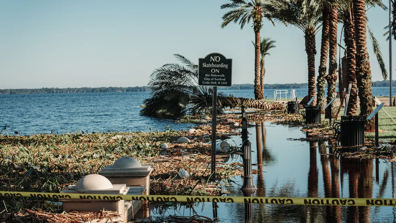 A Florida lakefront path after a storm: standing water, washed-up debris and caution tape between the palms