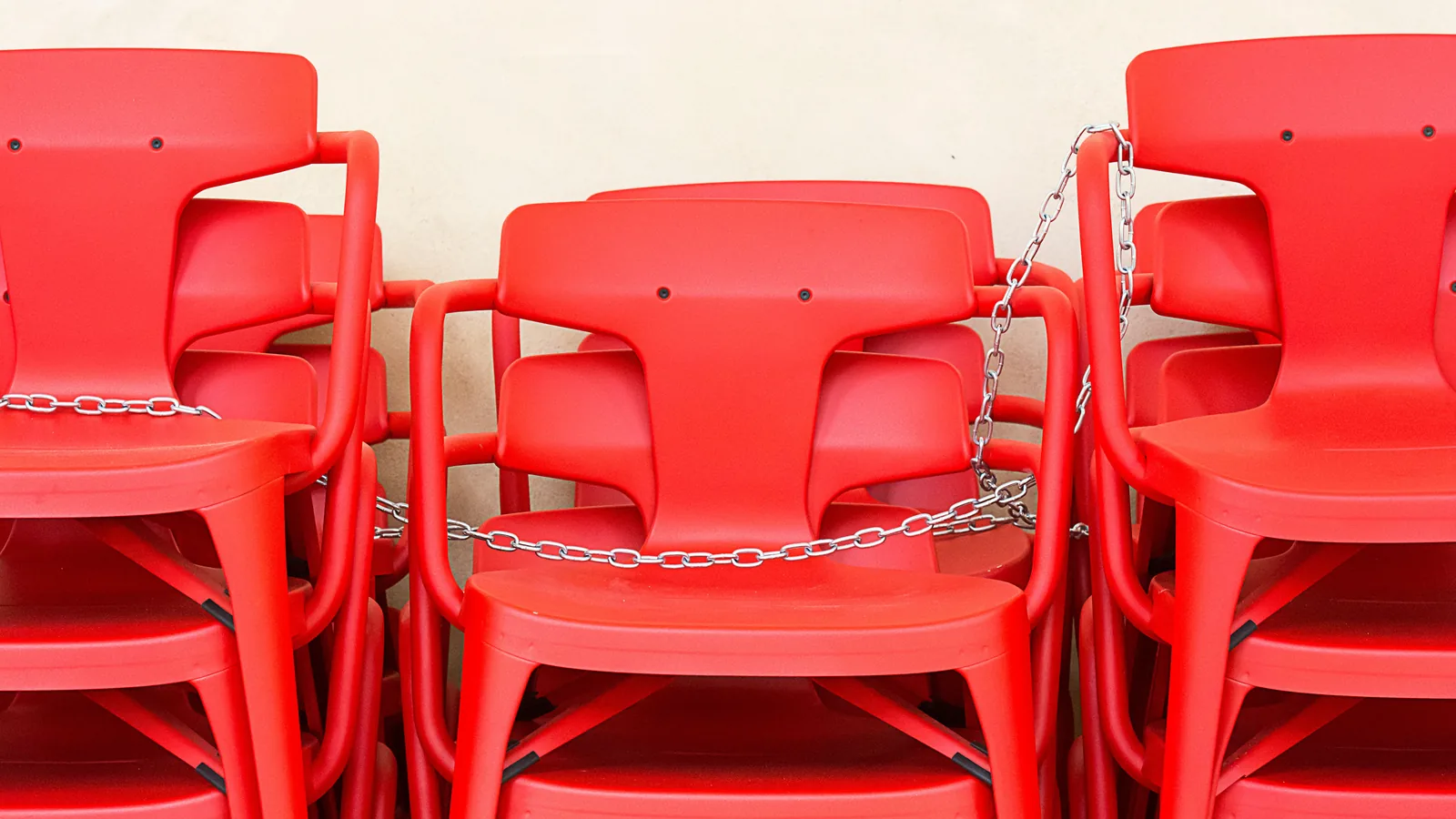 Stacks of red plastic chairs threaded together with a steel chain against a plain wall