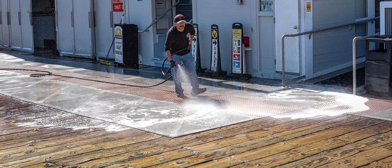 A worker pressure washing a walkway, with a sheet of soapy water spreading across the boards around him
