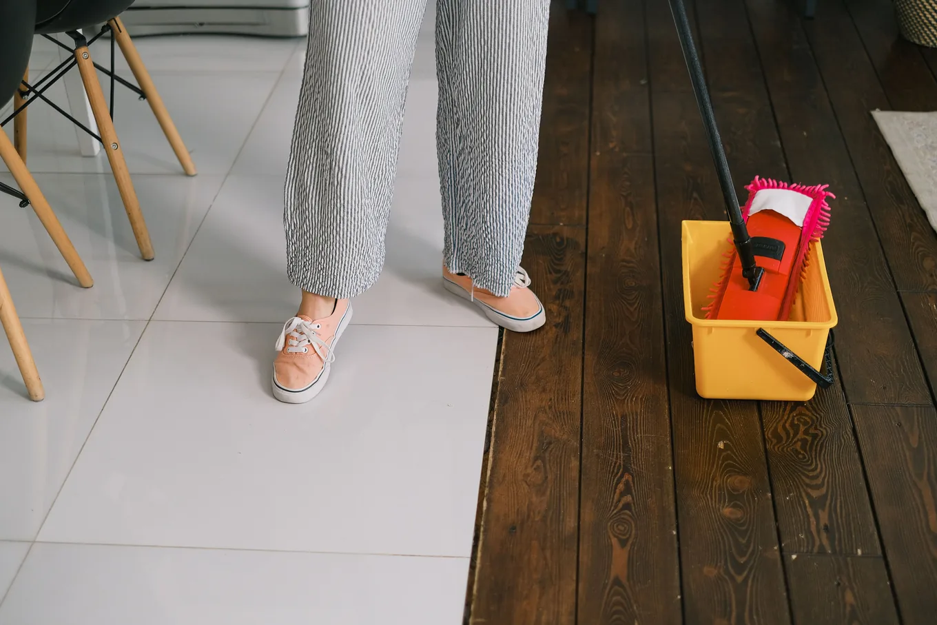 A flat microfiber mop dipped into a small yellow bucket on a floor, beside a person&rsquo;s sneakers