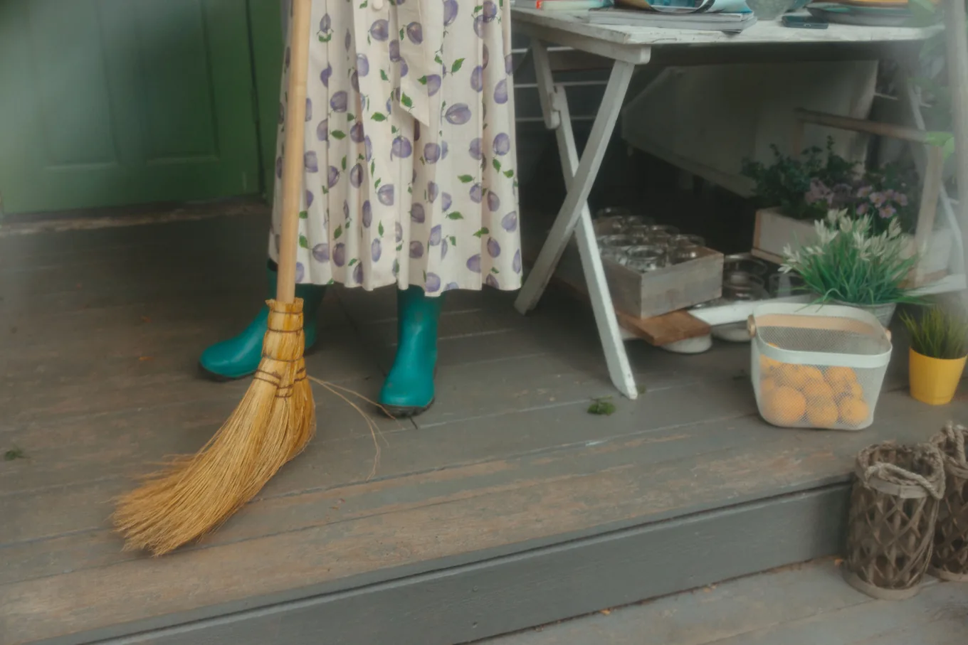 Someone in rubber boots sweeping a gray painted wooden porch floor with a straw broom, next to potted plants