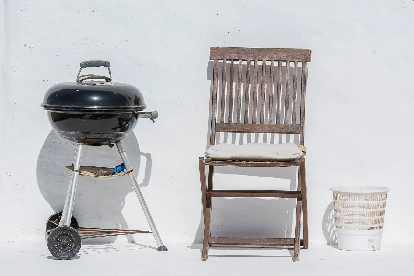 A charcoal kettle grill beside a folding wooden chair against a white wall outdoors