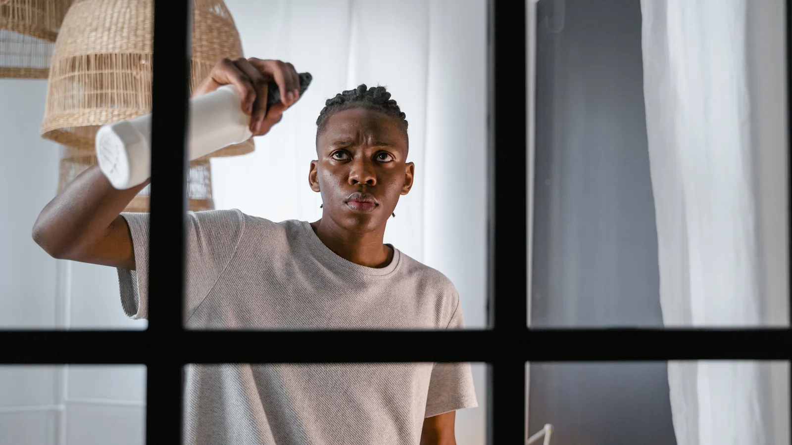 A man holding a spray bottle up to a tall window frame, mid-clean