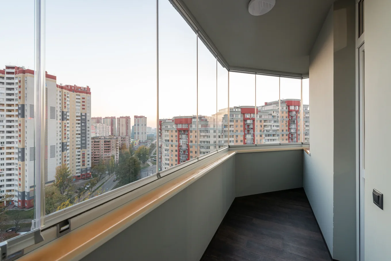 A narrow glazed balcony running along an apartment, floor-to-ceiling glass panels looking out over a city