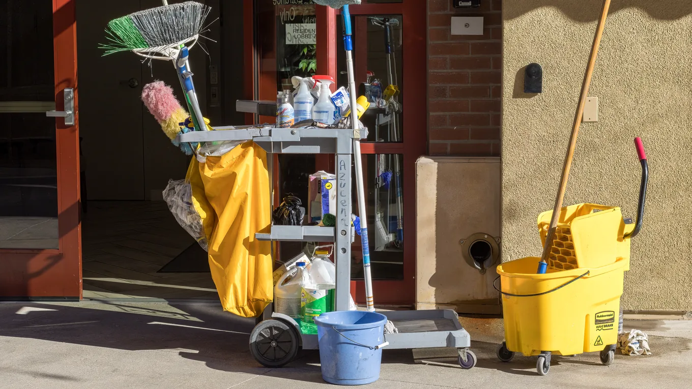 A loaded janitorial cart, mop bucket and long-handled tools standing outside a building entrance
