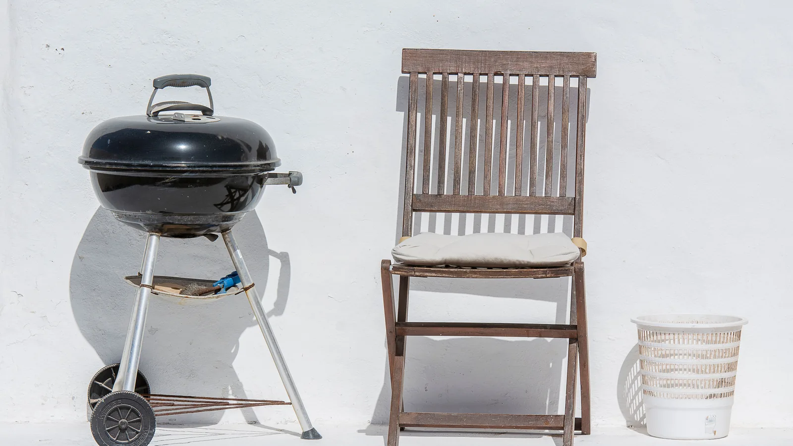 A charcoal kettle grill beside a wooden folding chair against a white wall