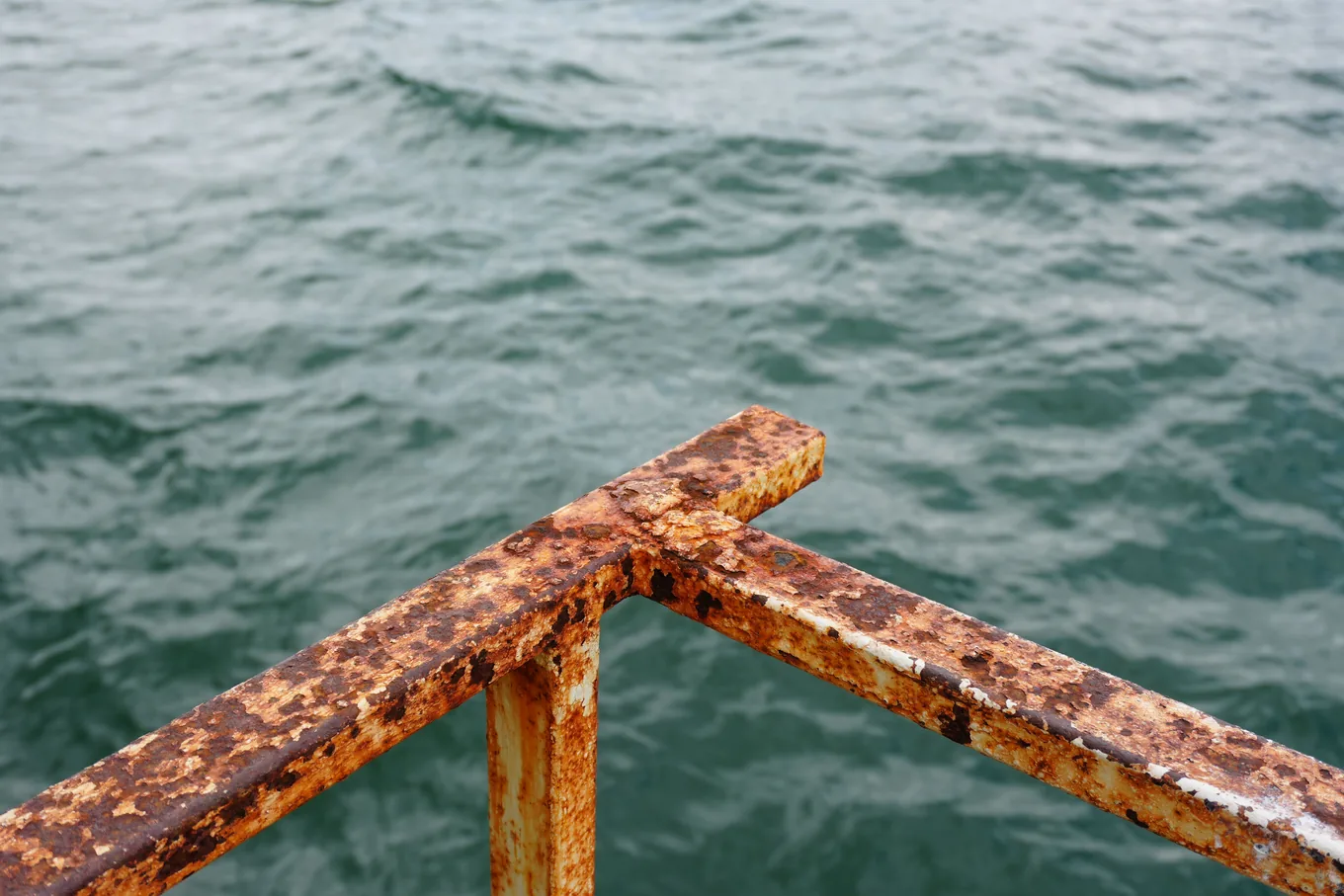 A corner of a painted steel railing, its paint flaking over orange rust, above choppy sea water