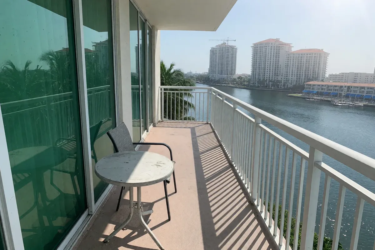 A condo balcony over the water: glass sliding doors on the left, a small table and chair, a white rail on the right.