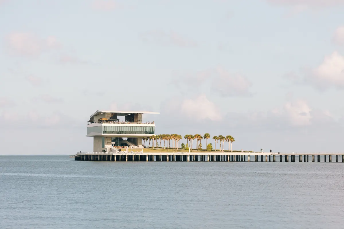St. Pete Pier head building with rooftop palms, sitting low on flat pale water under a soft sky