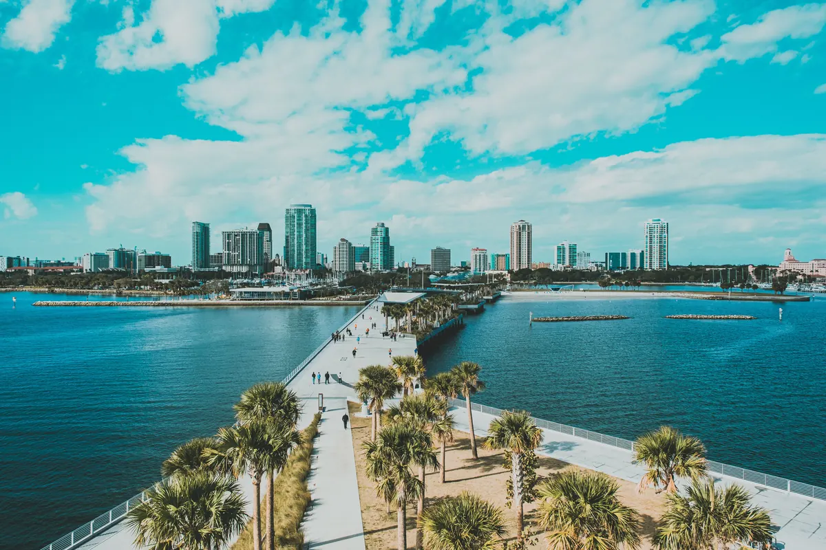 Palm-lined walkway of the St. Pete Pier leading toward the downtown St. Petersburg skyline across teal water